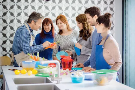 Tupperwareparty mit fünf Personen, die in einer Küche einem Vorsteller lauschen.