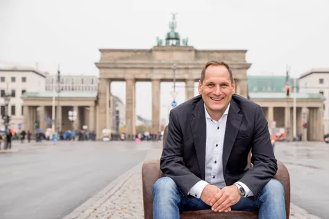 Hauptstadtcoach Oliver Zigann sitzt auf einem Sofa vor dem Brandenburger Tor und lacht in die Kamera.