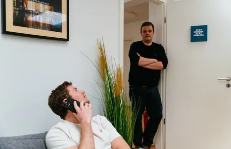 Axel telefoniert auf der Couch und schaut zu Christian der in der Tür steht.